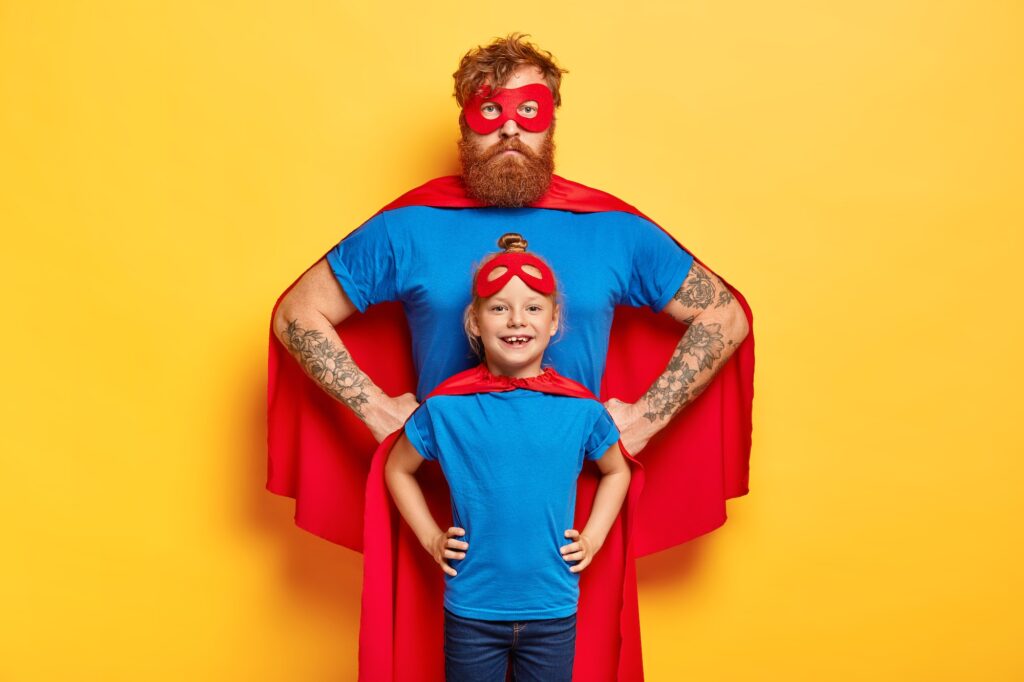 Photo of confident ginger father and daughter keep hands on waists, pretend being superhero and defe